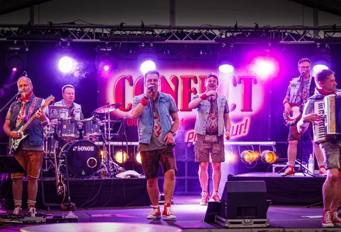 Six-member band performing live on stage under purple lights; vocalist center, other members on guitar, drums, and accordion, with a bright banner behind them.