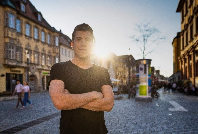 Man in a black t-shirt stands with arms crossed in a cobblestone town square, sun glare behind him and historic buildings on both sides.