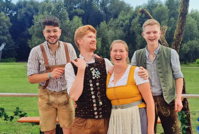 Four friends outdoors in a park, wearing traditional Bavarian clothing (lederhosen and dirndl), smiling and posing together.